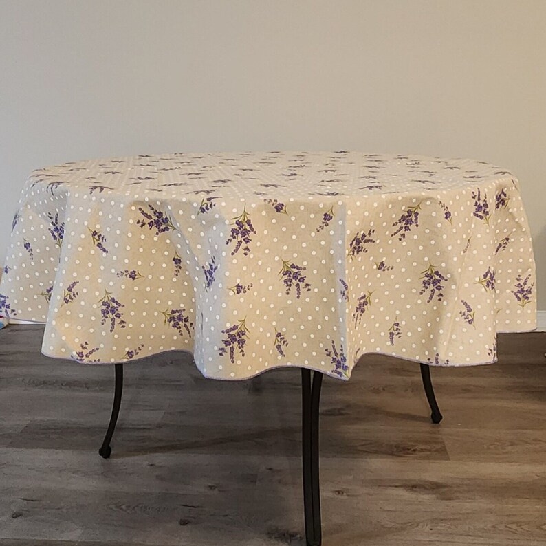 May include: Round beige tablecloth with a pattern of white polka dots and purple lavender sprigs. The tablecloth is draped over a black metal table with four legs. The fabric has a scalloped edge.