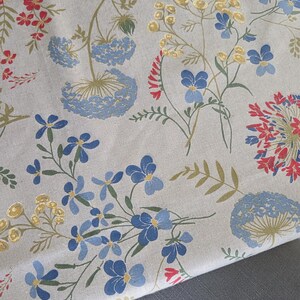 May include: Floral fabric featuring a repeating pattern of blue, red, and yellow flowers with green stems and leaves on a beige background. The fabric is folded over a gray surface, showcasing the pattern's design.