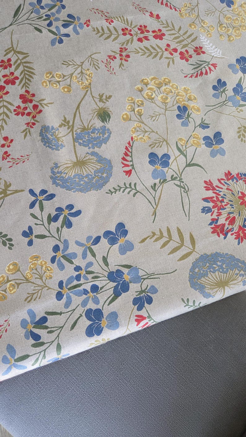 May include: Floral fabric featuring a repeating pattern of blue, red, and yellow flowers with green stems and leaves on a beige background. The fabric is folded over a gray surface, showcasing the pattern's design.