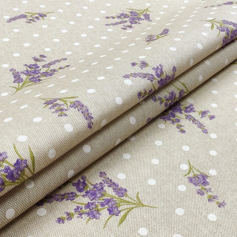 May include: Beige fabric with a repeating pattern of purple lavender sprigs and white polka dots. The fabric is folded, showing the texture and design. The lavender has green stems and leaves.