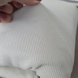 May include: Close-up of a light gray textured fabric, possibly a pillow cover or upholstery material. The fabric has a woven pattern with a subtle diagonal texture. The material is being held up, revealing its thickness and construction.