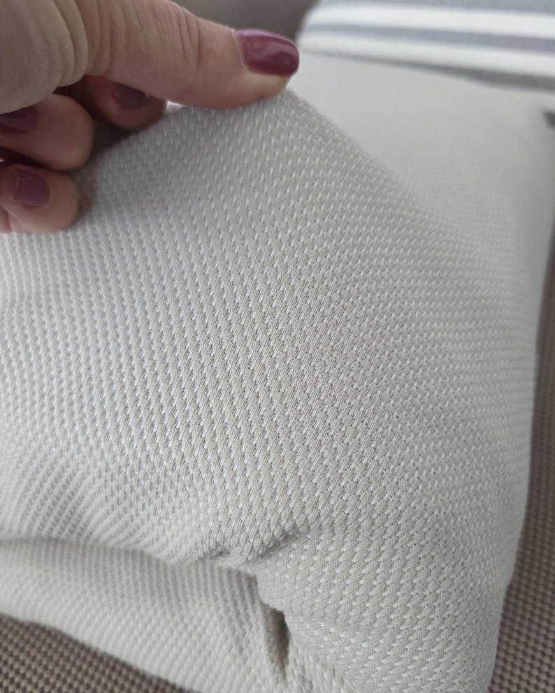 May include: Close-up of a light gray textured fabric, possibly a pillow cover or upholstery material. The fabric has a woven pattern with a subtle diagonal texture. The material is being held up, revealing its thickness and construction.