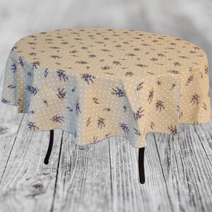 May include: A round beige tablecloth with a pattern of white polka dots and purple lavender sprigs. The tablecloth is draped over a small, round table with black legs. The overall aesthetic is rustic and floral.