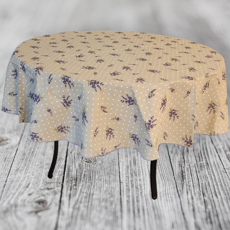 May include: A round beige tablecloth with a pattern of white polka dots and purple lavender sprigs. The tablecloth is draped over a small, round table with black legs. The overall aesthetic is rustic and floral.
