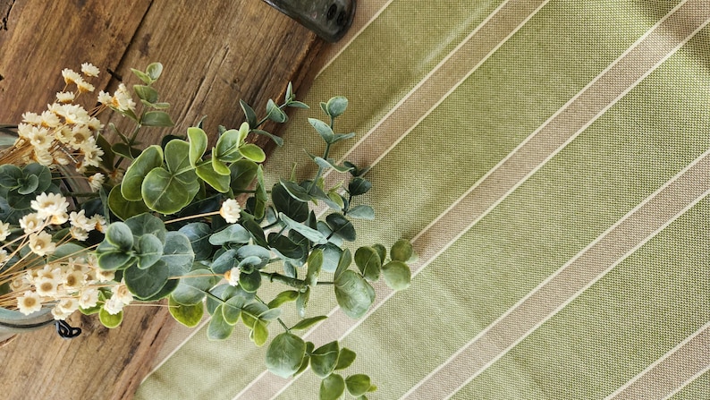 K&ouml;nnte beinhalten: Nahaufnahme einer gr&uuml;n-wei&szlig; gestreiften Tischdecke mit einem Strau&szlig; getrockneter wei&szlig;er Blumen und gr&uuml;nen Eukalyptusbl&auml;ttern.