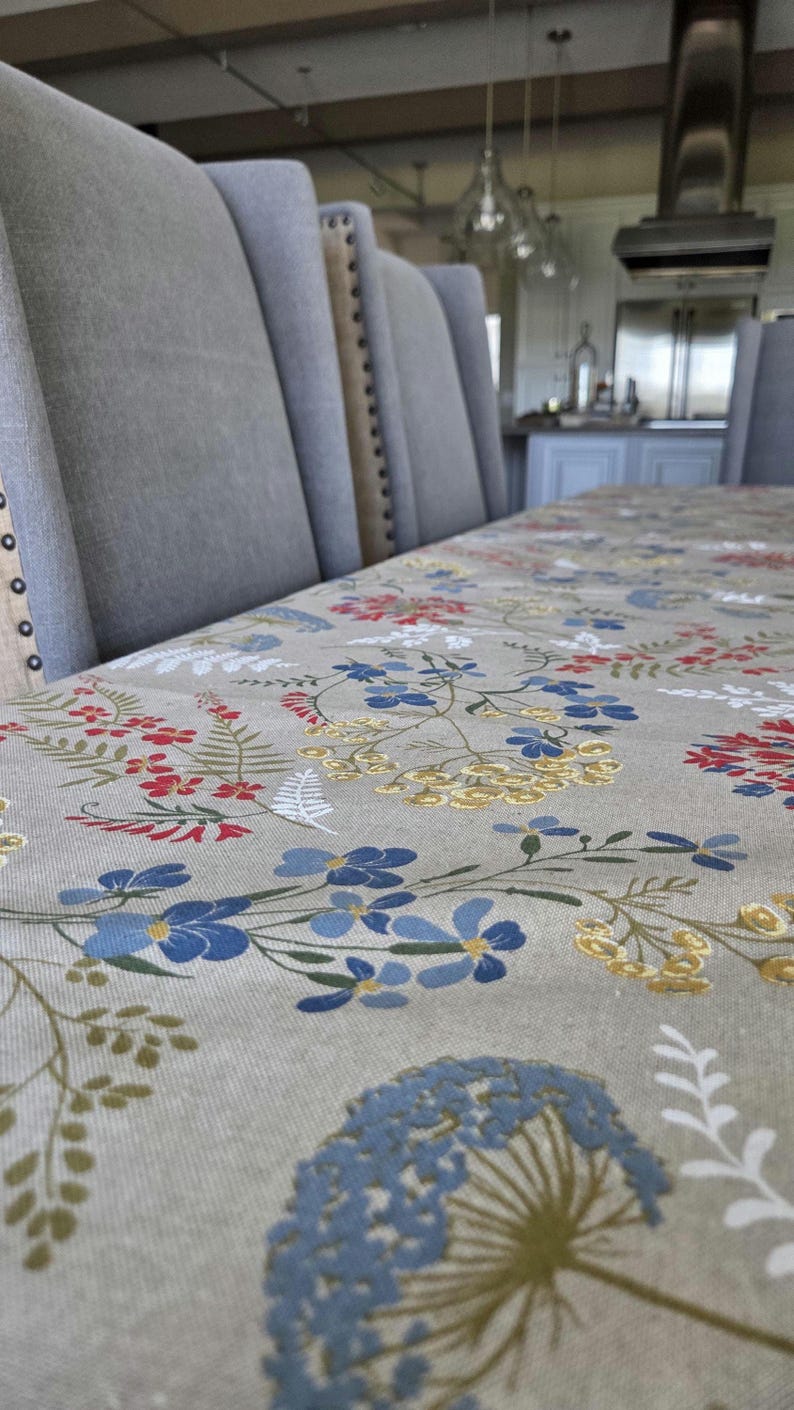 May include: A dining table set with upholstered gray chairs and a floral tablecloth. The tablecloth features a pattern of red, blue, and yellow flowers on a neutral background. The chairs have button detailing.
