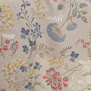 May include: Close-up of a beige fabric with a floral pattern. The design features blue, yellow, and red flowers with green stems and leaves. The flowers are scattered across the fabric.
