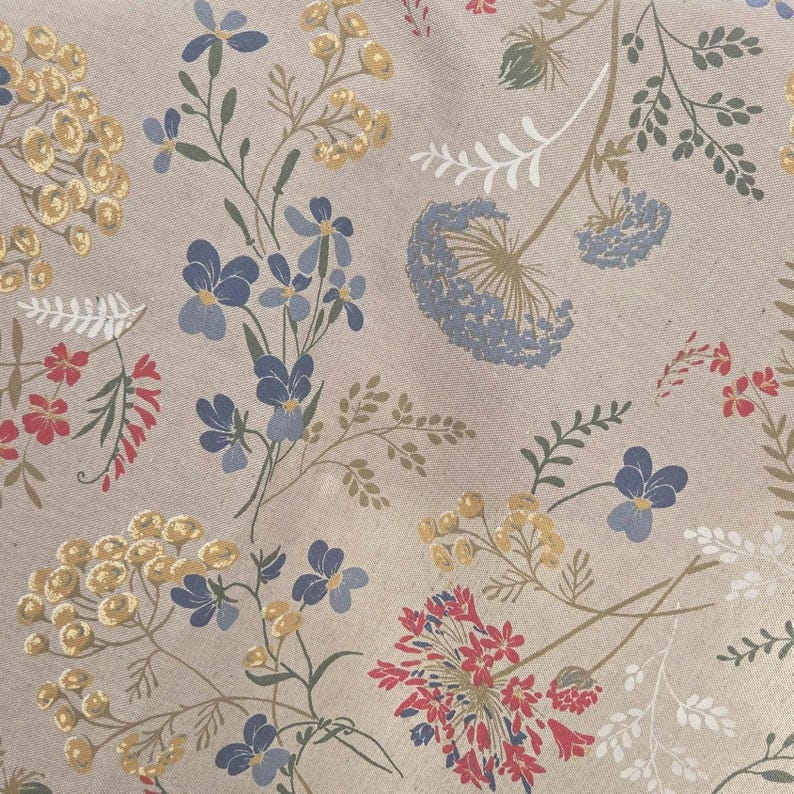 May include: Close-up of a beige fabric with a floral pattern. The design features blue, yellow, and red flowers with green stems and leaves. The flowers are scattered across the fabric.