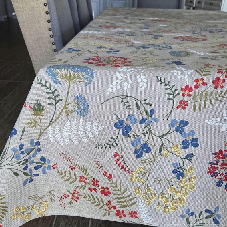 May include: A beige tablecloth with a floral pattern featuring blue, red, and yellow flowers and green leaves. The tablecloth is draped over a table, with a chair visible in the background. The floral design adds a touch of nature to the dining setting.