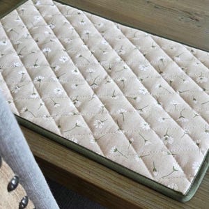May include: A quilted placemat featuring a beige fabric adorned with a repeating pattern of small white flowers and green stems. The placemat has a green border and is placed on a wooden surface.
