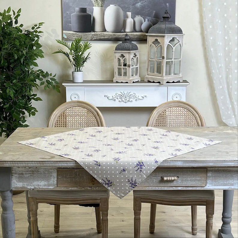 May include: A gray and white patterned table runner with lavender floral accents and white polka dots. The runner is placed on a rustic wooden table. Decorative lanterns and vases are in the background.