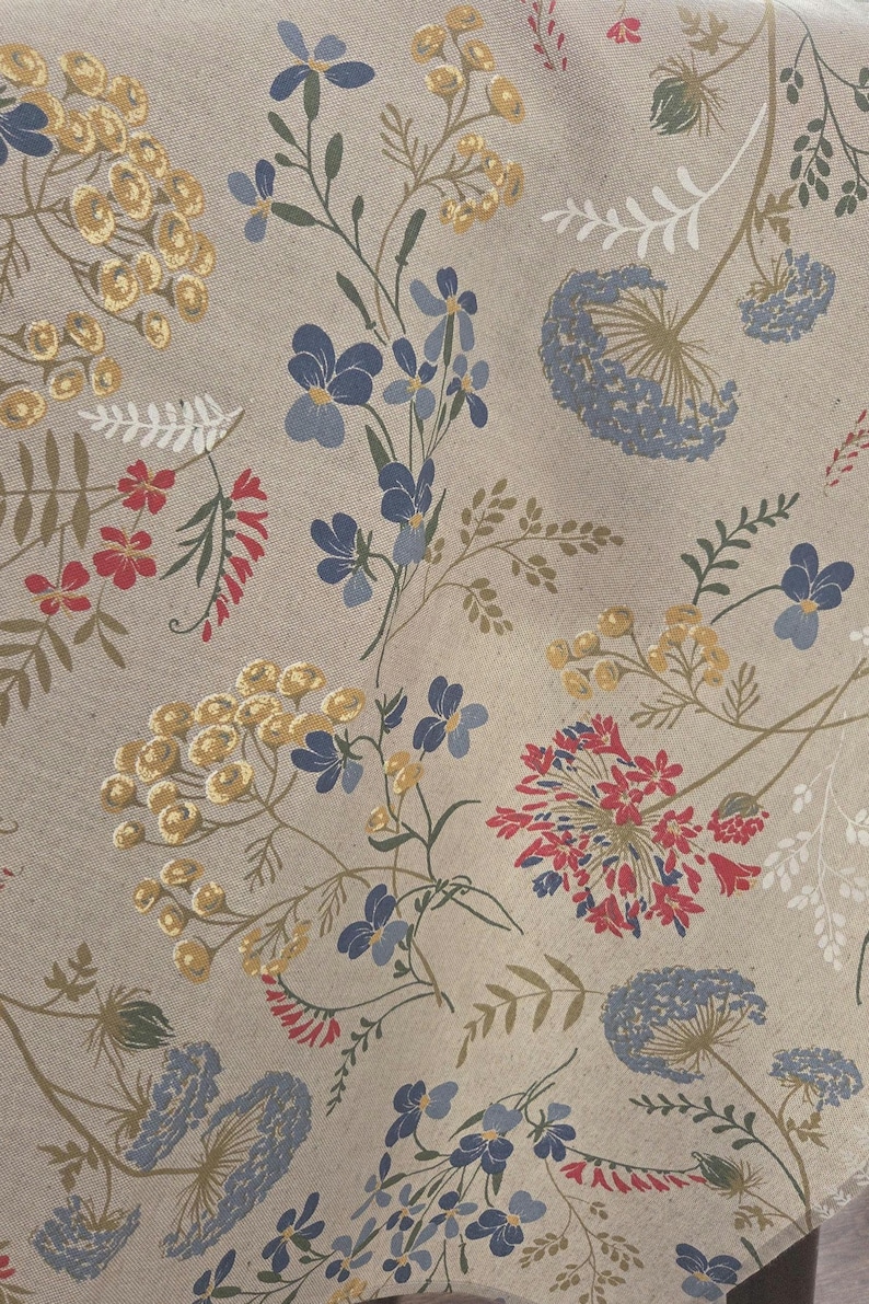 May include: A beige fabric with a floral pattern featuring blue, yellow, and red flowers. The design includes various flower types and green stems, creating a natural, botanical theme. The fabric is likely used for home decor or textiles.