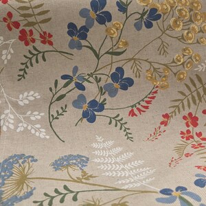 May include: Close-up of a beige fabric with a floral pattern. The design features blue, red, and yellow flowers, along with green and white foliage. The fabric has a natural, textured appearance.
