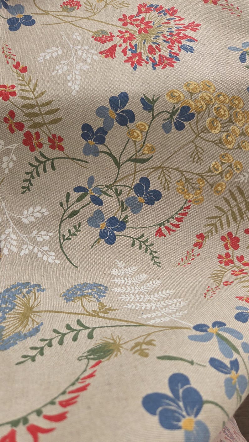 May include: Close-up of a beige fabric with a floral pattern. The design features blue, red, and yellow flowers, along with green and white foliage. The fabric has a natural, textured appearance.