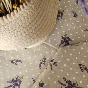May include: A woven basket with dried wheat and yellow flowers sits on a beige fabric. The fabric is patterned with white polka dots and sprigs of purple lavender, creating a rustic, floral theme.