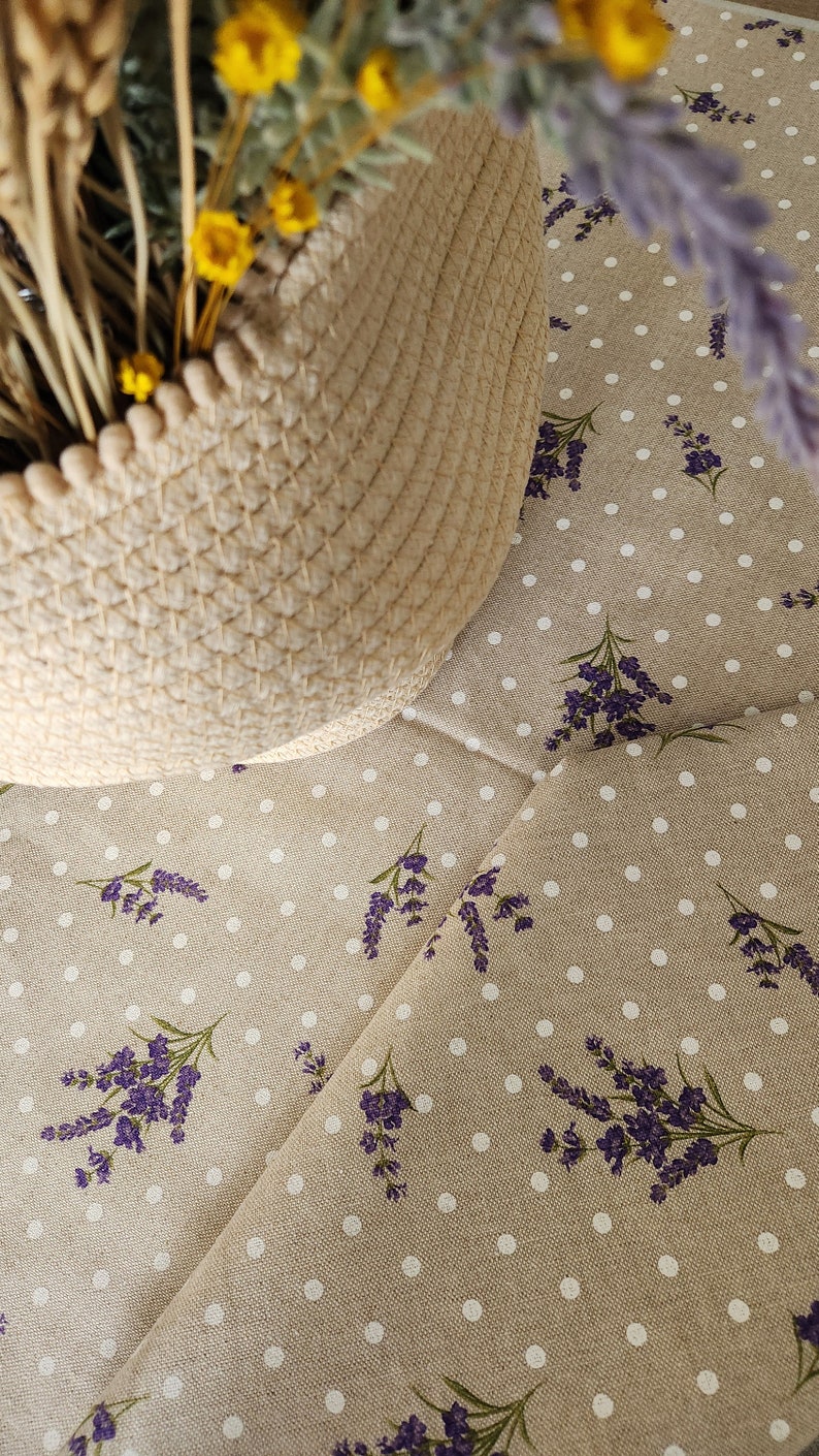 May include: A woven basket with dried wheat and yellow flowers sits on a beige fabric. The fabric is patterned with white polka dots and sprigs of purple lavender, creating a rustic, floral theme.