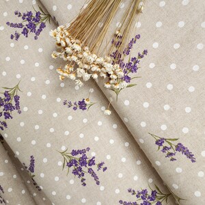 May include: Beige fabric with a pattern of white polka dots and purple lavender sprigs. Dried flowers are arranged on top of the fabric. The fabric is folded, showing the pattern and texture.