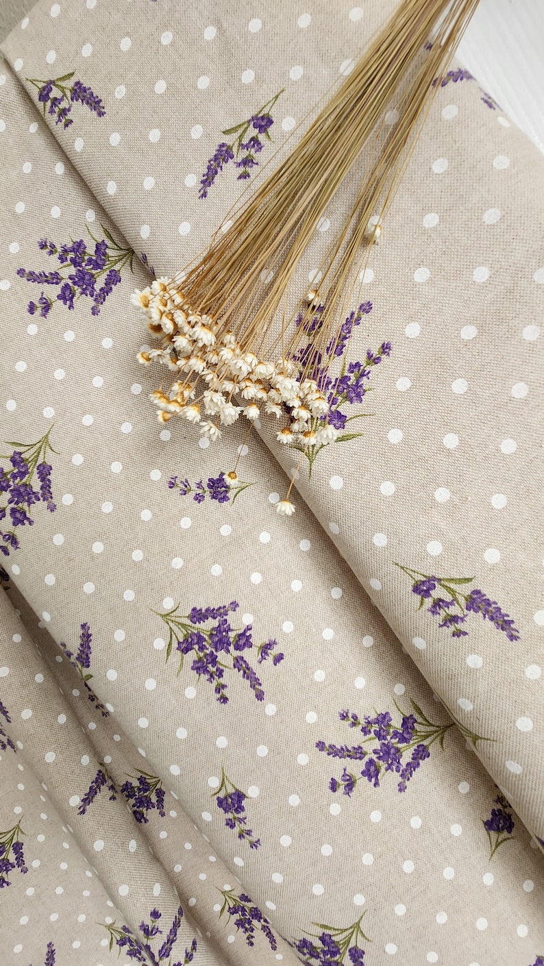 May include: Beige fabric with a pattern of white polka dots and purple lavender sprigs. Dried flowers are arranged on top of the fabric. The fabric is folded, showing the pattern and texture.
