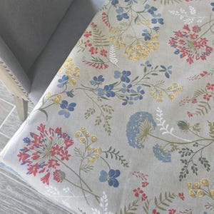 May include: A floral patterned fabric featuring a variety of flowers in red, blue, and yellow against a neutral background. The fabric is draped over a chair, showcasing the design's potential use in home decor.
