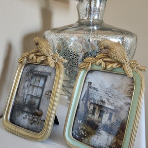 May include: Two ornate picture frames with gold trim and bird accents. The frames are on a white surface and feature vintage-style images of houses.