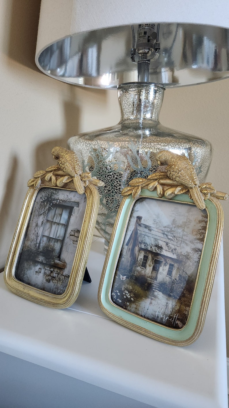 May include: Two ornate picture frames with gold trim and bird accents. The frames are on a white surface and feature vintage-style images of houses.