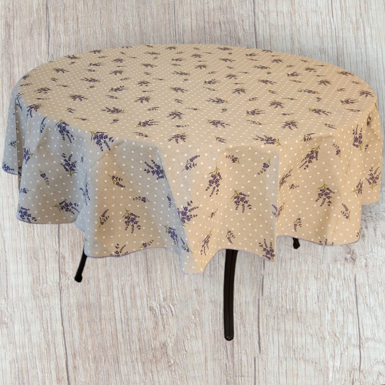 May include: A round tablecloth with a beige background, white polka dots, and purple lavender sprigs. The tablecloth is draped over a table with black legs. The fabric has a natural, slightly textured appearance.