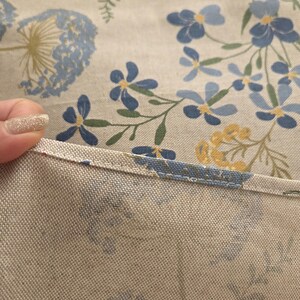 May include: Close-up of a floral patterned fabric. The fabric features a light beige background with blue, yellow, and green floral designs. The edge of the fabric is visible, showing a stitched seam.