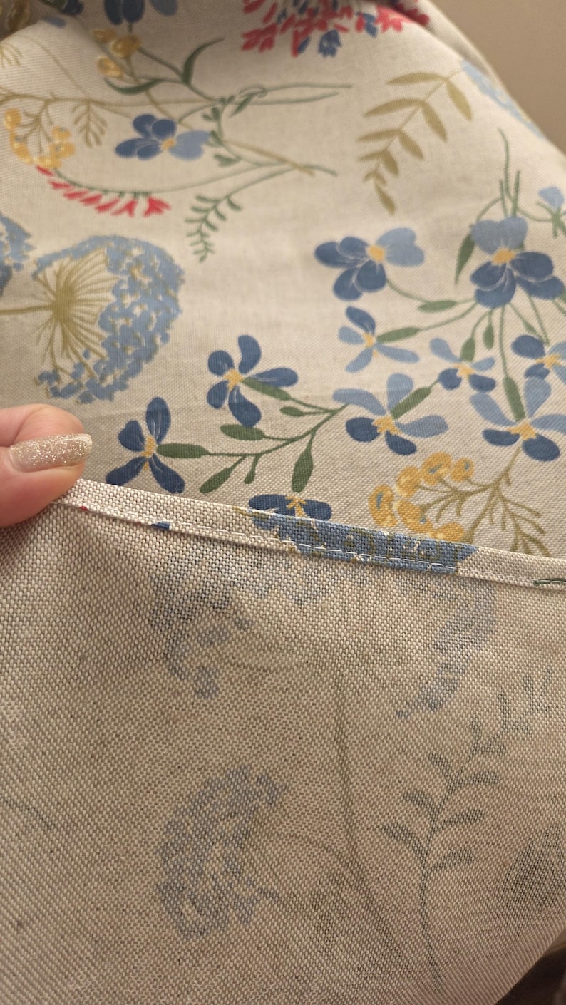 May include: Close-up of a floral patterned fabric. The fabric features a light beige background with blue, yellow, and green floral designs. The edge of the fabric is visible, showing a stitched seam.