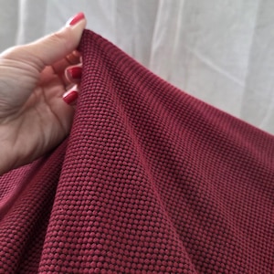 May include: Close-up of a textured, burgundy fabric. The fabric has a raised, dotted pattern, creating a unique visual and tactile experience. The material is held by a hand with red nail polish, showcasing the fabric's drape and texture.