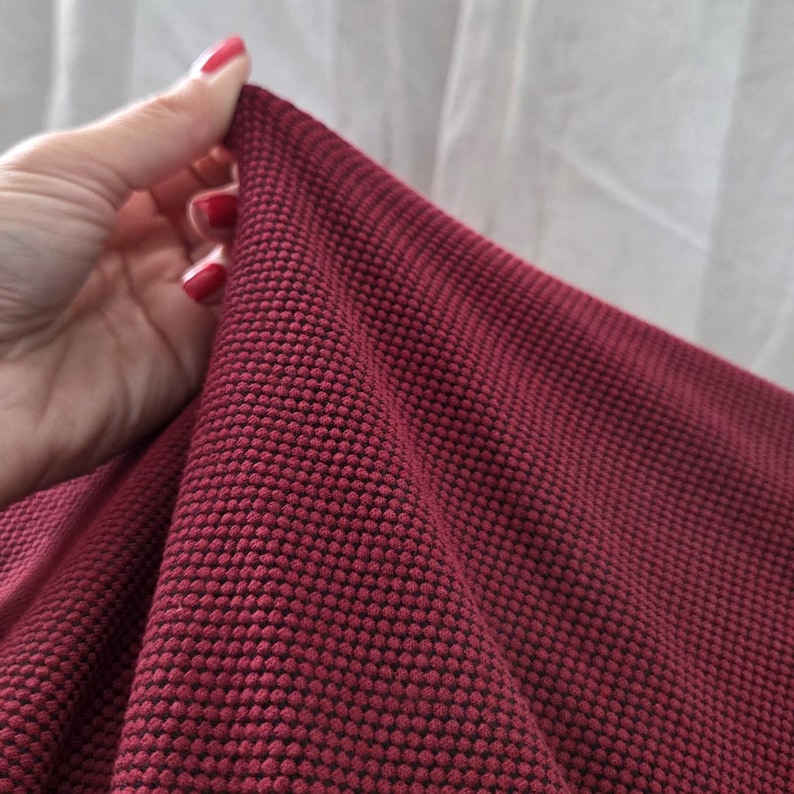 May include: Close-up of a textured, burgundy fabric. The fabric has a raised, dotted pattern, creating a unique visual and tactile experience. The material is held by a hand with red nail polish, showcasing the fabric's drape and texture.