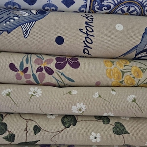 May include: A stack of patterned fabric rolls. The top roll features a blue and white geometric design. Below, rolls display floral and botanical patterns in various colors, including purple, yellow, and white. One roll has the word "profondo" printed on it.