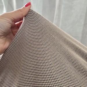 May include: Close-up of a beige fabric with a repeating pattern of small, raised dots. The fabric is held up, revealing its texture and the contrast between the beige dots and the darker background.