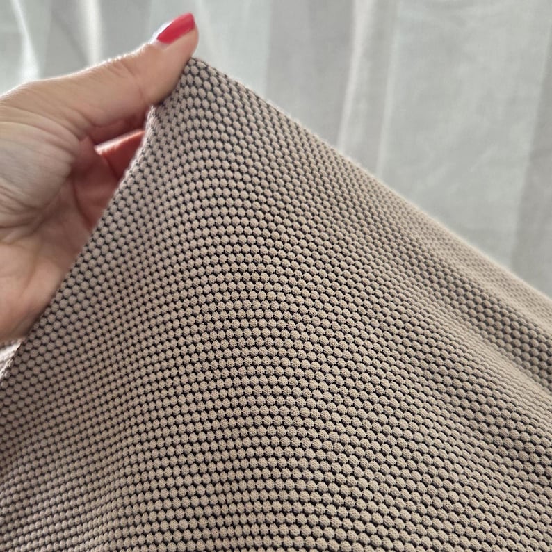 May include: Close-up of a beige fabric with a repeating pattern of small, raised dots. The fabric is held up, revealing its texture and the contrast between the beige dots and the darker background.
