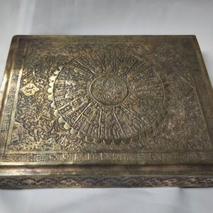 Puede incluir: Una caja cuadrada de latón con intrincados diseños florales y geométricos. La parte superior de la caja presenta un diseño circular con un motivo central.