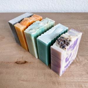 May include: Five bars of handmade soap in various colors and scents. The bars are arranged in a row from left to right, with the lavender soap on the right. The soaps are all rectangular and have a slightly textured surface.