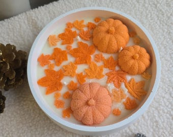 Könnte beinhalten: Ein runder weißer Behälter gefüllt mit weißem Wachs, verziert mit drei orangefarbenen kürbisförmigen Wachsstücken und zahlreichen kleinen orangefarbenen Ahornblatt-Wachsakzenten auf der Oberfläche der Kerze.