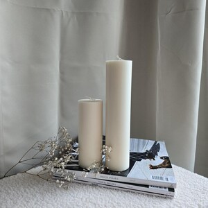 Peut inclure: Deux bougies blanches en forme de pilier de tailles diff&eacute;rentes sur une pile de magazines avec un petit bouquet de fleurs blanches.