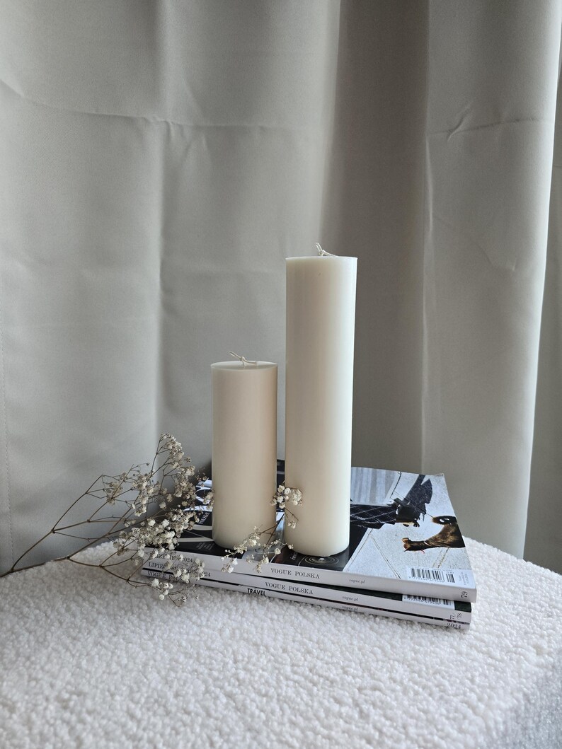 Peut inclure: Deux bougies blanches en forme de pilier de tailles diff&eacute;rentes sur une pile de magazines avec un petit bouquet de fleurs blanches.