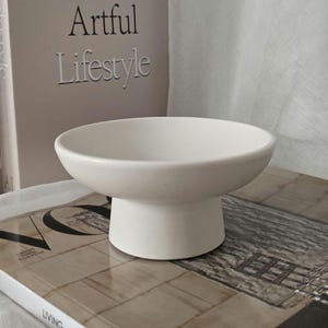 Peut inclure: Un bol en céramique blanc mat doté d'un bassin large et peu profond ainsi que d'une base cylindrique épaisse. Le bol présente une surface lisse et unie avec un léger mouchetage, offrant une apparence propre et uniforme pour ce récipient.