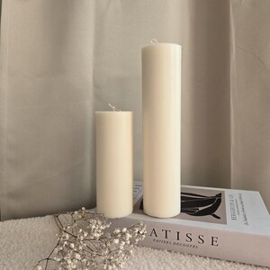 Peut inclure: Deux bougies blanches en forme de pilier de tailles diff&eacute;rentes sur une surface blanche avec un livre intitul&eacute; "ATISSE PAPIERS DECOUPES" et un petit bouquet de fleurs blanches.