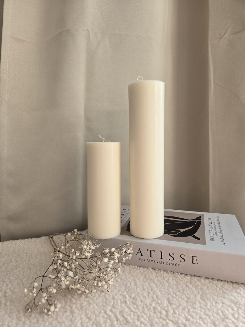 Peut inclure: Deux bougies blanches en forme de pilier de tailles diff&eacute;rentes sur une surface blanche avec un livre intitul&eacute; "ATISSE PAPIERS DECOUPES" et un petit bouquet de fleurs blanches.