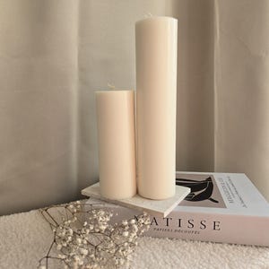 Peut inclure: Deux bougies blanches en forme de pilier de tailles diff&eacute;rentes sur une surface textur&eacute;e blanche avec un livre intitul&eacute; "ATISSE PAPIERS DECOUPES" et une brindille de fleurs blanches.