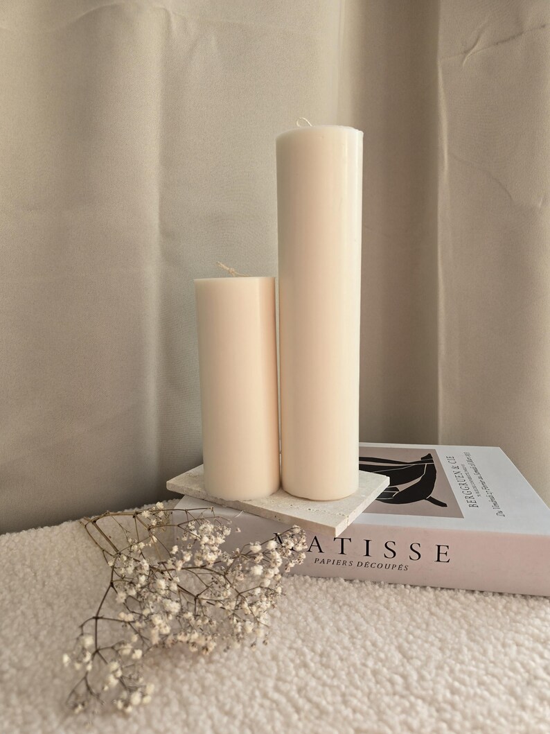 Peut inclure: Deux bougies blanches en forme de pilier de tailles diff&eacute;rentes sur une surface textur&eacute;e blanche avec un livre intitul&eacute; "ATISSE PAPIERS DECOUPES" et une brindille de fleurs blanches.