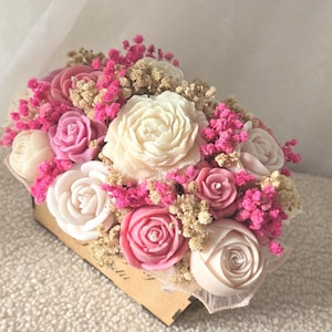 Puede incluir: Una caja rectangular de madera llena de un arreglo de velas con forma de rosas blancas y rosas, intercaladas con racimos de flores secas de paniculata en tonos rosa y beige. Las velas están dispuestas en un grupo denso y superpuesto.