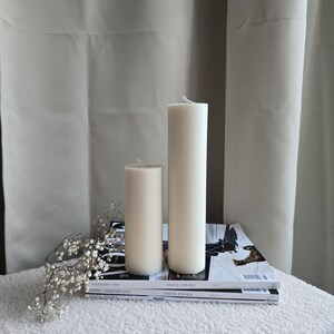 Peut inclure: Deux bougies blanches en forme de pilier de tailles diff&eacute;rentes sur une surface moelleuse blanche avec une pile de magazines Vogue Polska.
