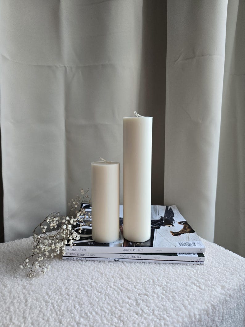 Peut inclure: Deux bougies blanches en forme de pilier de tailles diff&eacute;rentes sur une surface moelleuse blanche avec une pile de magazines Vogue Polska.
