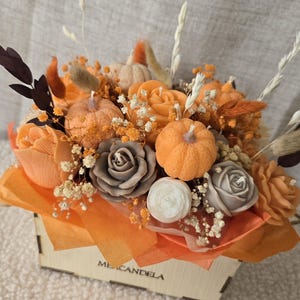 Może przedstawiać: Drewniana skrzynka z napisem ME CANDELA zawierająca kompozycję świec w kształcie pomarańczowych i szarych r&oacute;ż, pomarańczowych dyń oraz małej białej świecy, ozdobiona suszonymi kwiatami, kłosami pszenicy i pomarańczową bibułą.