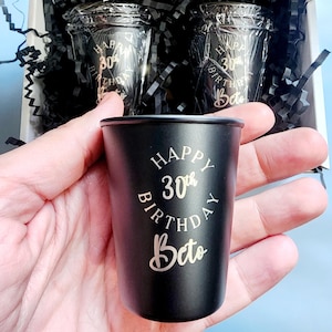 May include: Two black shot glasses with gold lettering that reads "Happy 30th Birthday Beto" in a circle. The shot glasses are in a box with black shredded paper.