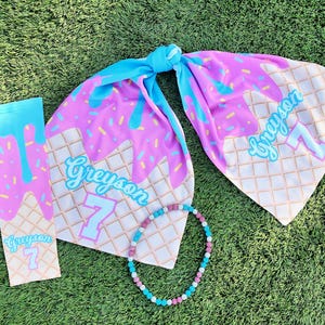 May include: A set of ice cream themed party supplies featuring a blue and pink ice cream cone design with the name Greyson and the number 7. The set includes a scarf, a sleeve, and a beaded necklace.