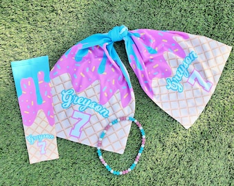 May include: A set of ice cream themed party supplies featuring a blue and pink ice cream cone design with the name Greyson and the number 7. The set includes a scarf, a sleeve, and a beaded necklace.